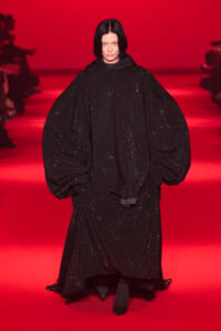 Model wearing an oversized black sequined cape on a red runway, mid-walk at a fashion show.