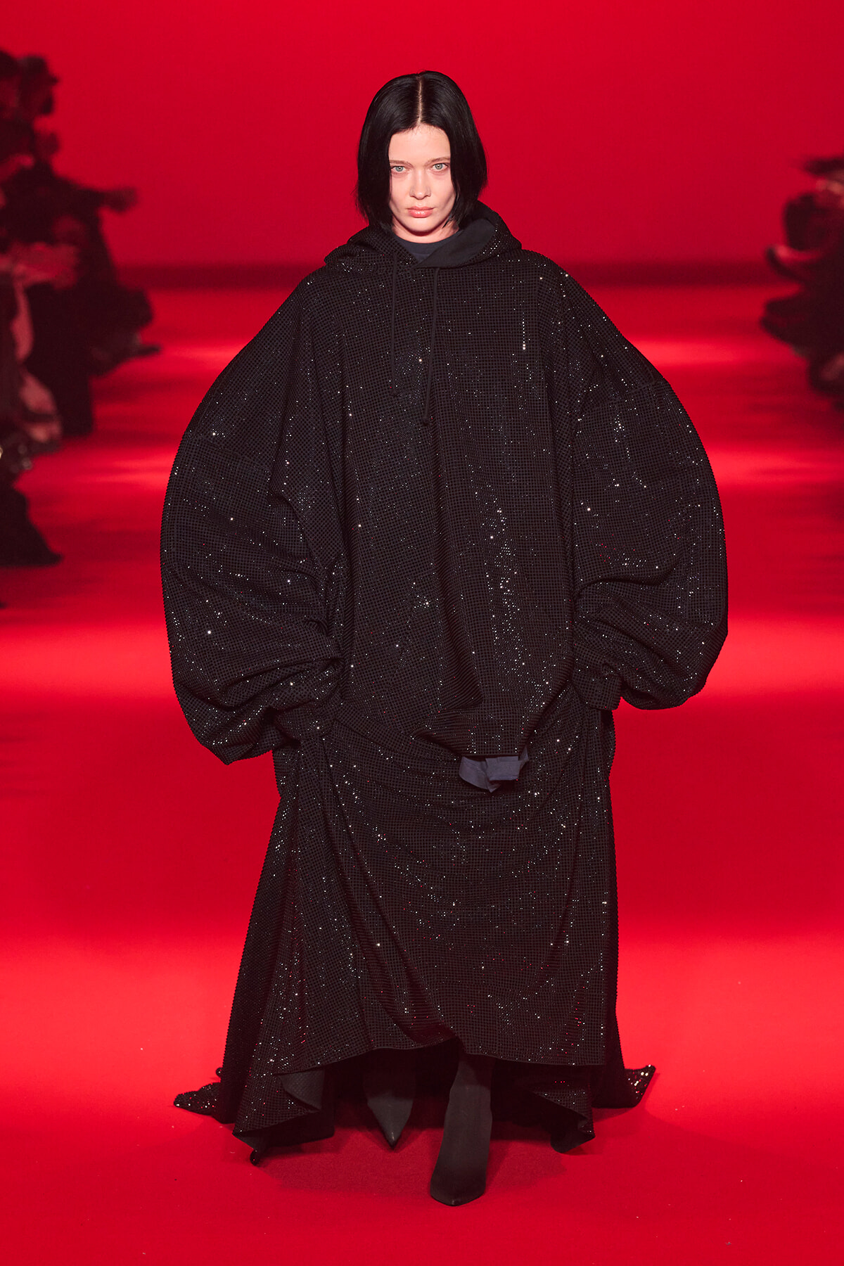 Model wearing an oversized black sequined cape on a red runway, mid-walk at a fashion show.