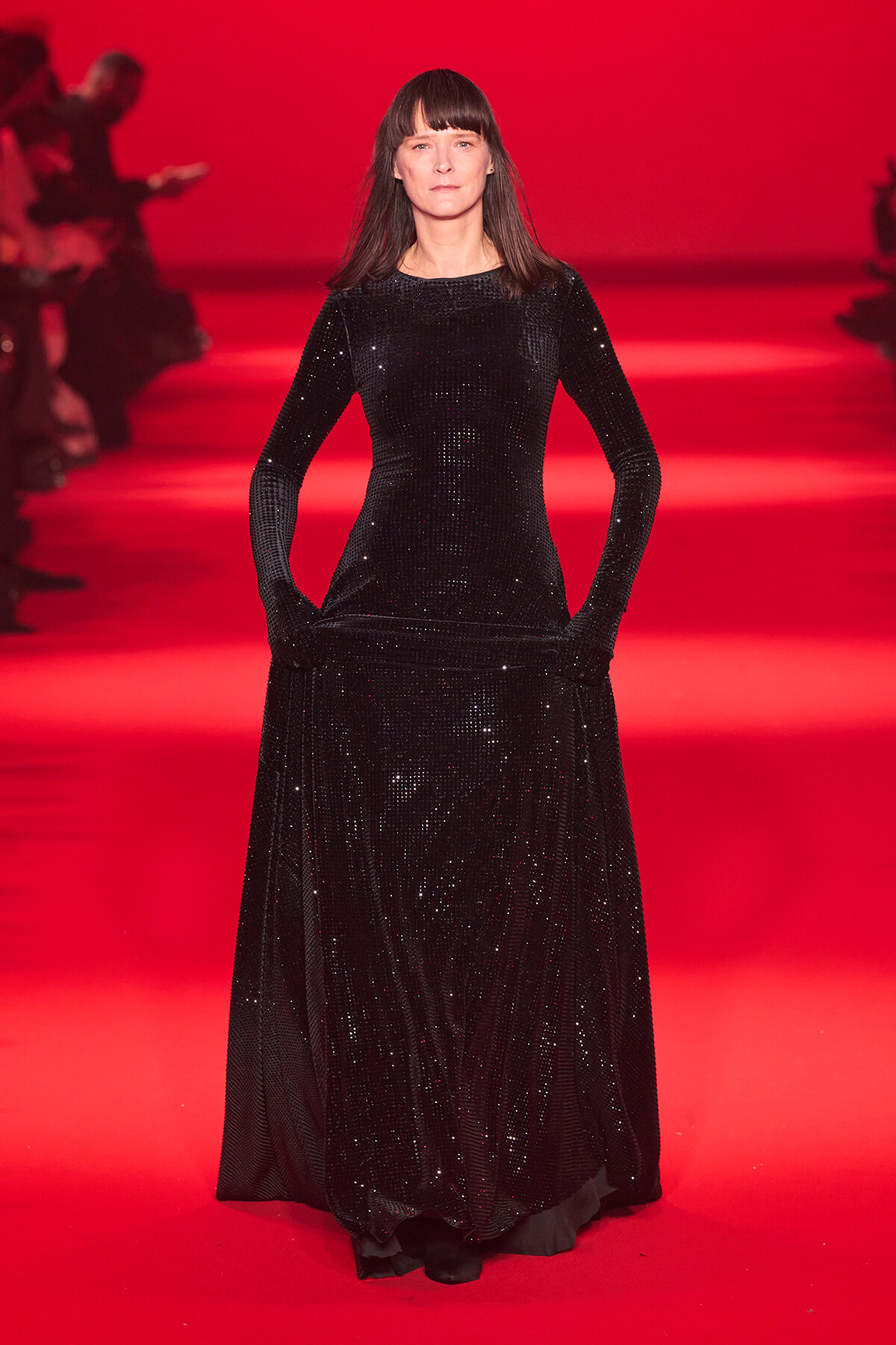 Model wearing a long black sequined gown on a red runway, hands in pockets, facing the camera.
