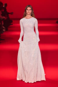 Model walks the runway in a long, shimmering white sequined gown against a red backdrop.