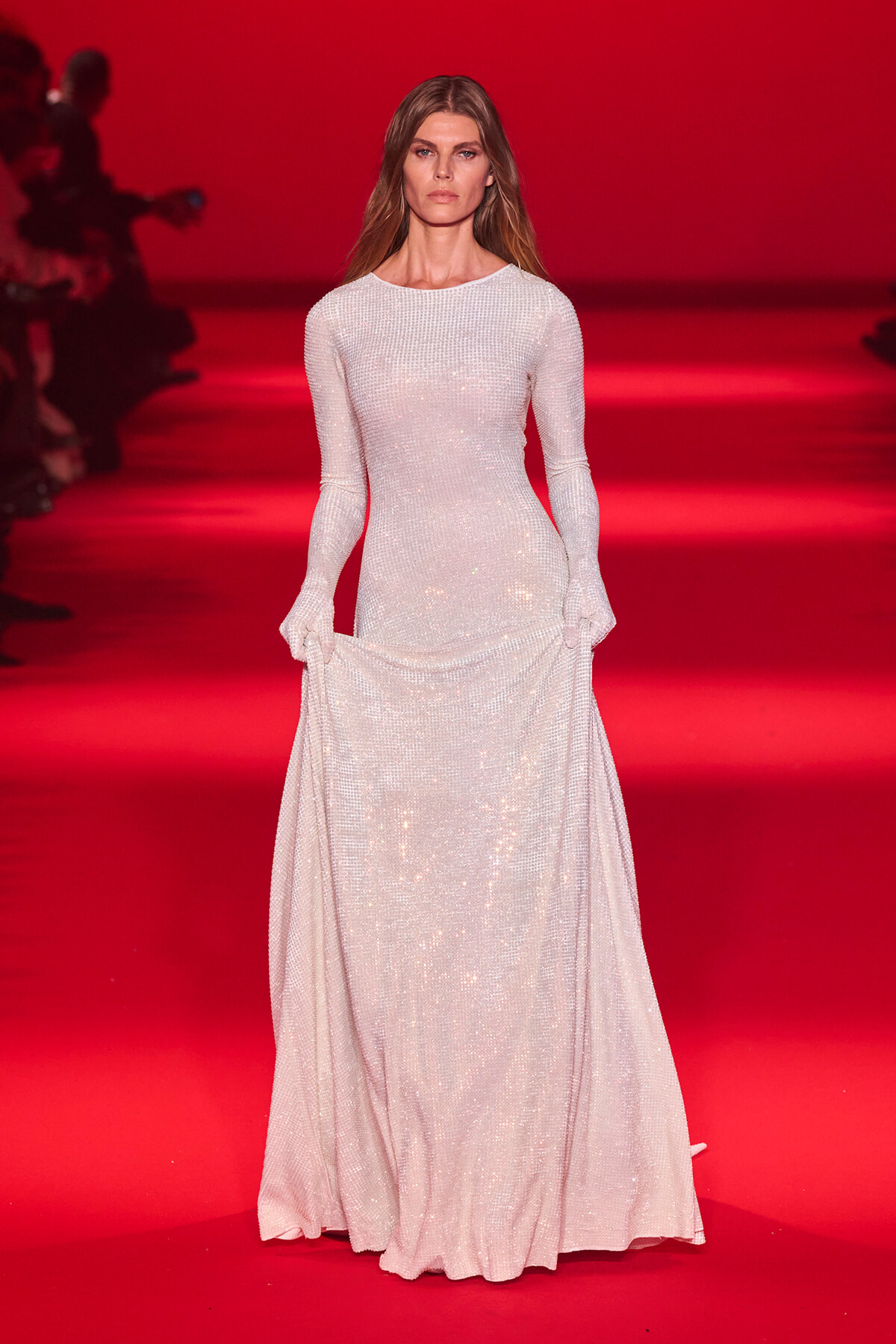 Model walks the runway in a long, shimmering white sequined gown against a red backdrop.