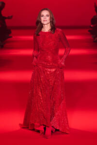 Woman wearing a floor-length red sequined gown on a red-lit runway, posing for a fashion show.