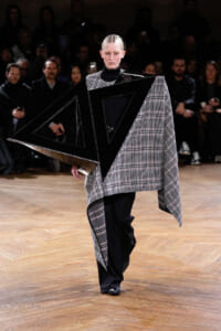 Model walks a fashion runway in a plaid cape with bold black geometric panels overlaying the garment.