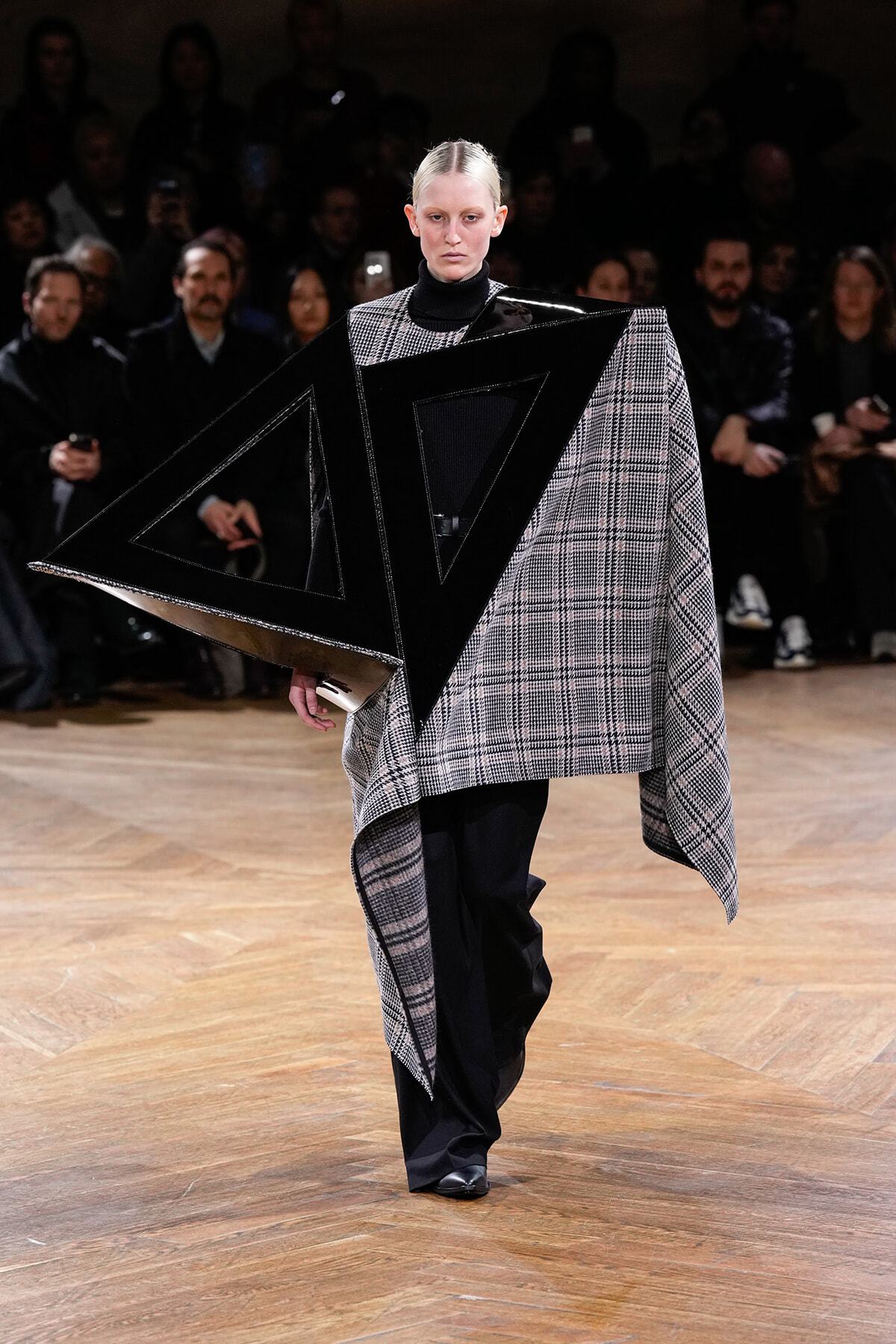 Model walks a fashion runway in a plaid cape with bold black geometric panels overlaying the garment.