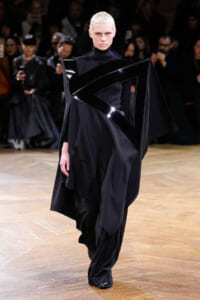Model on a runway wearing an avant-garde black ensemble with a glossy angular sash across the chest and shoulders; dark cape and high-neck top.