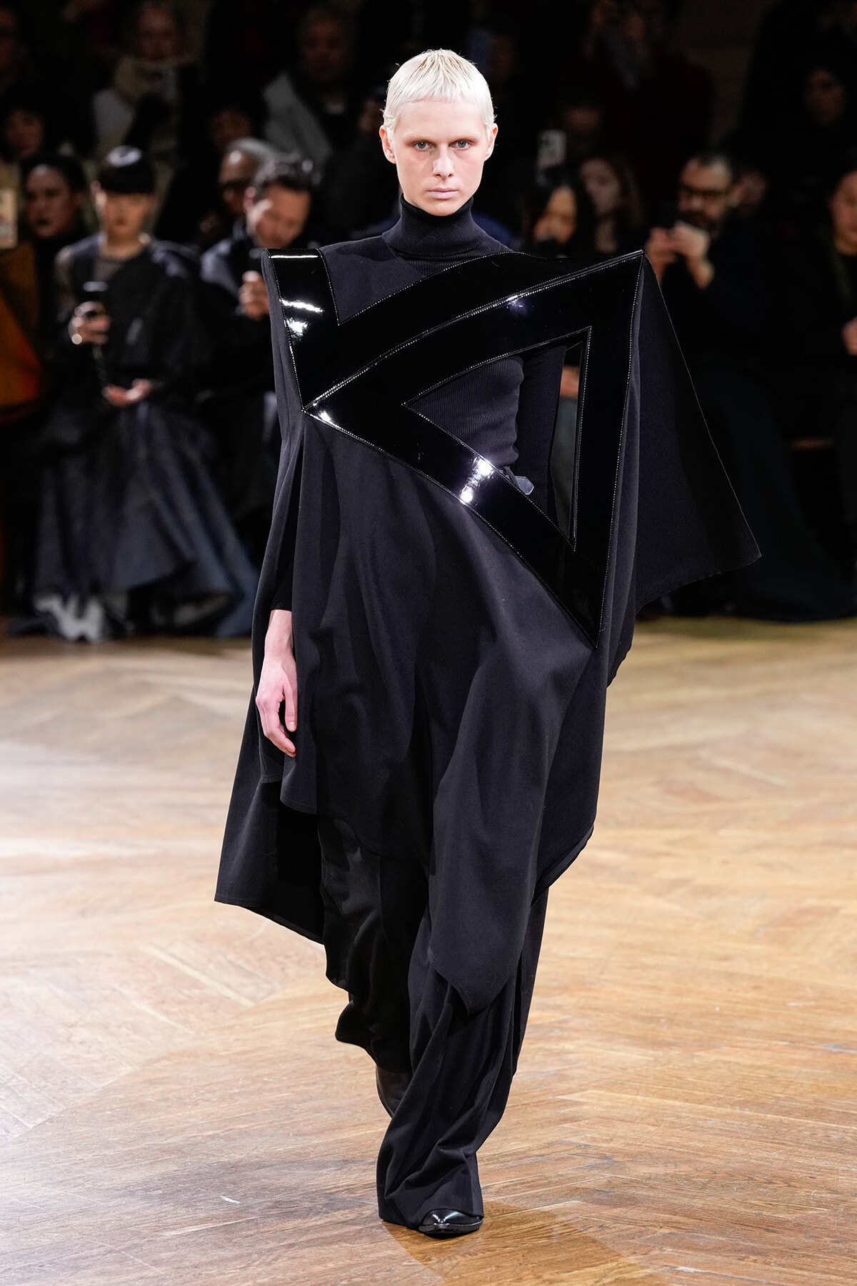 Model on a runway wearing an avant-garde black ensemble with a glossy angular sash across the chest and shoulders; dark cape and high-neck top.