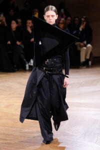 Model walks a runway wearing an all-black avant-garde outfit with a large geometric black triangle accessory over the torso.