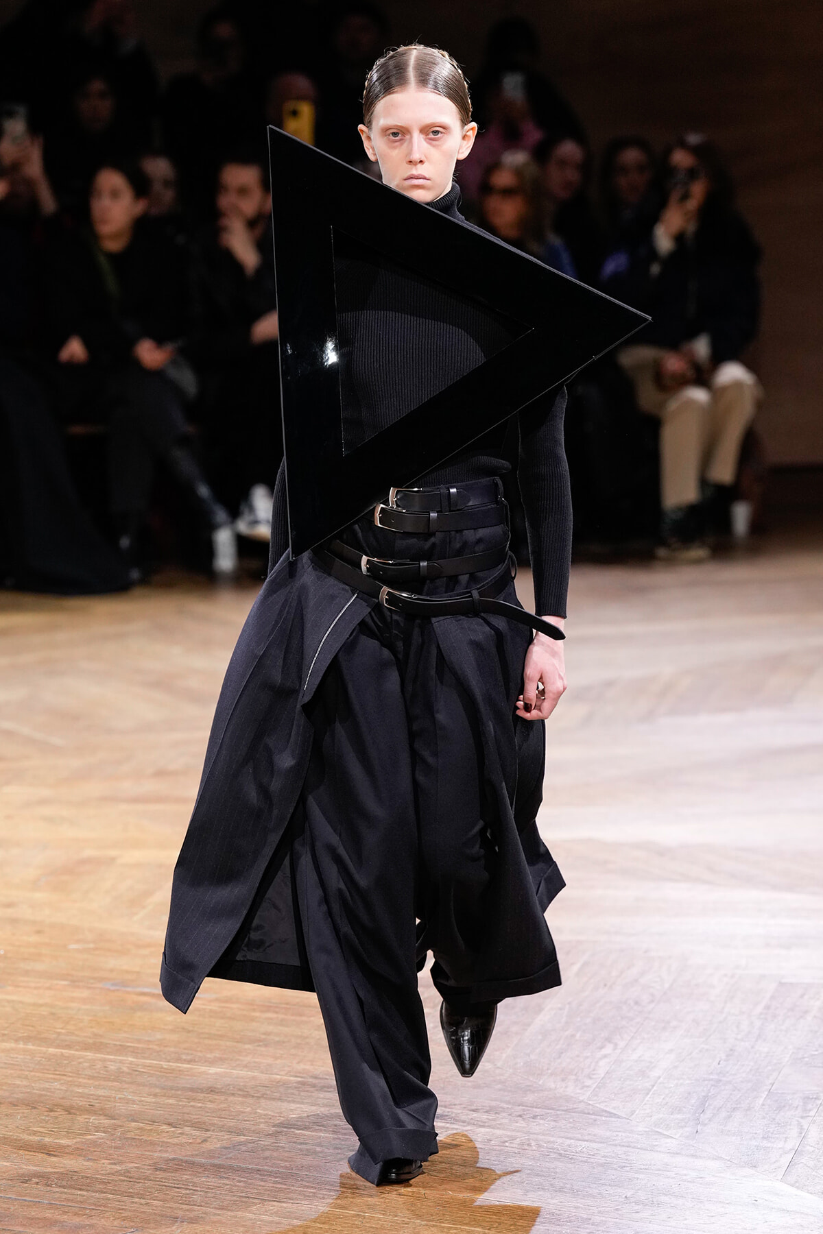 Model walks a runway wearing an all-black avant-garde outfit with a large geometric black triangle accessory over the torso.