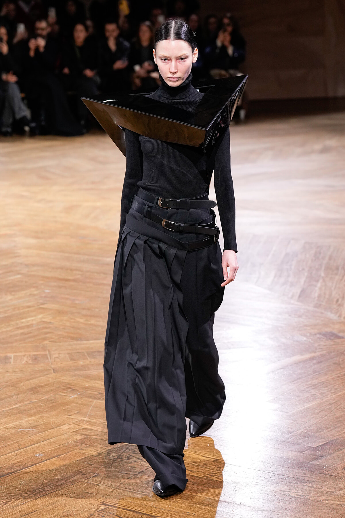Runway model in a black outfit with a large glossy angular collar extending over the shoulders.