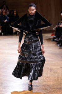Runway model in a futuristic black ensemble with a large geometric angular shoulder piece and a belted pleated skirt.