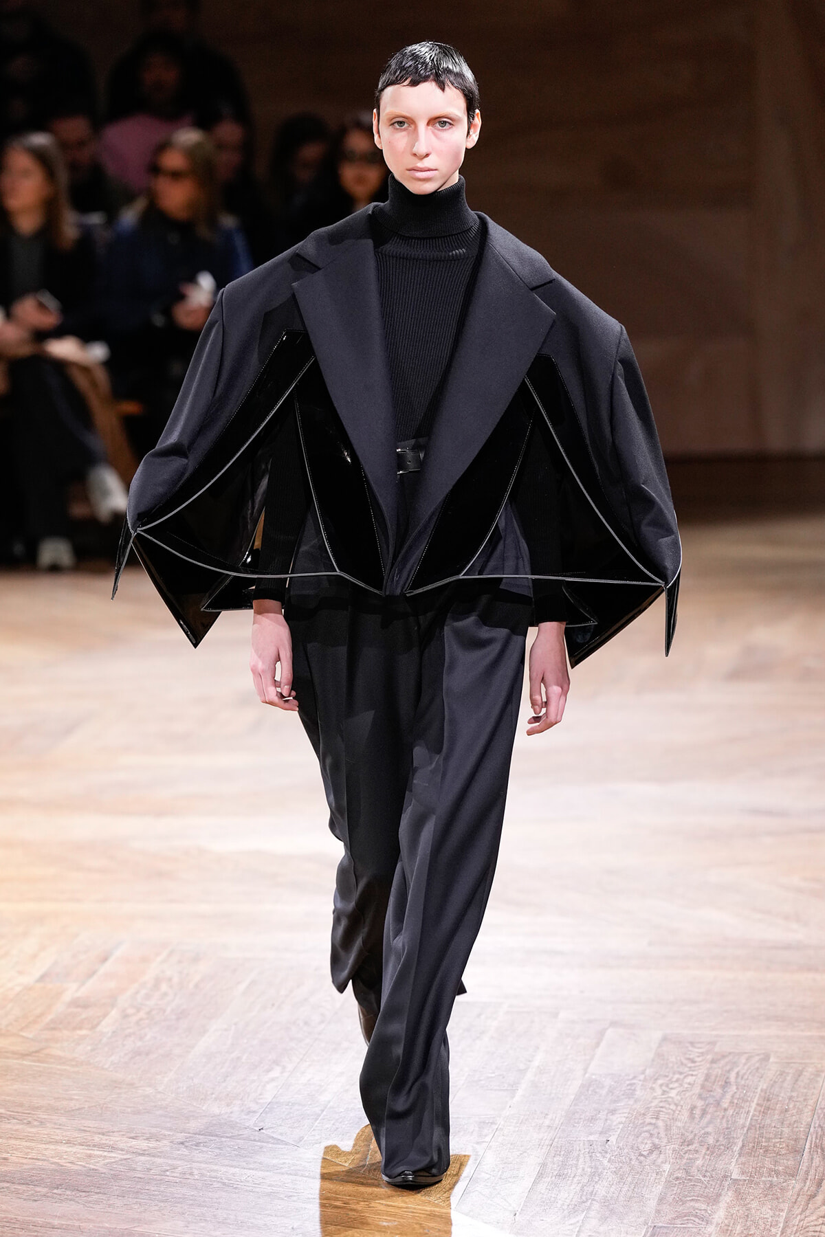 Model on a runway wearing an oversized black cape-like coat with angular panels and a black turtleneck, walking toward the camera.