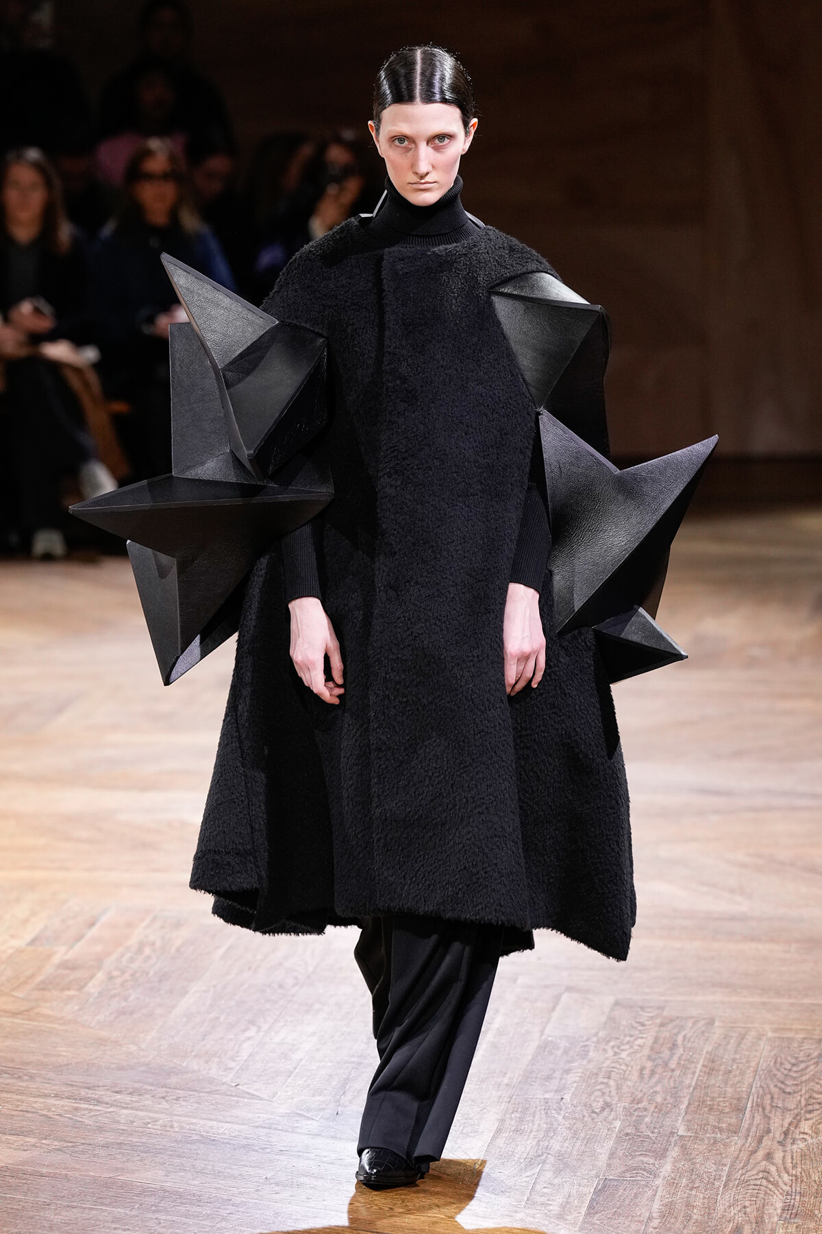 Runway model wearing a dramatic black textured coat with large geometric angular shoulder sculptures, walking on a wooden runway.