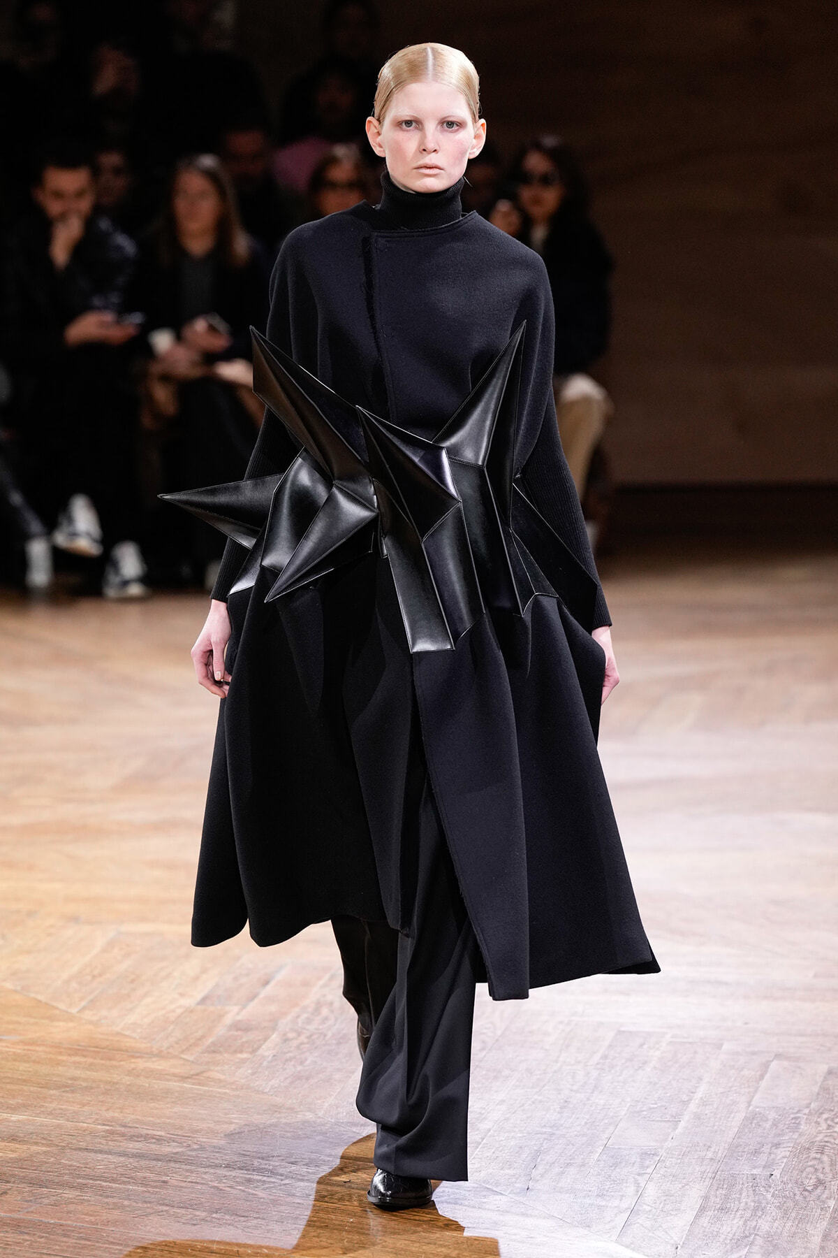 Model on a runway wearing an all-black outfit with a large sculptural leather star across the torso/waist area05 (avant-garde design).