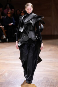 Model walking the runway in a futuristic black armor-inspired outfit with sculptural layered panels and oversized shoulders.