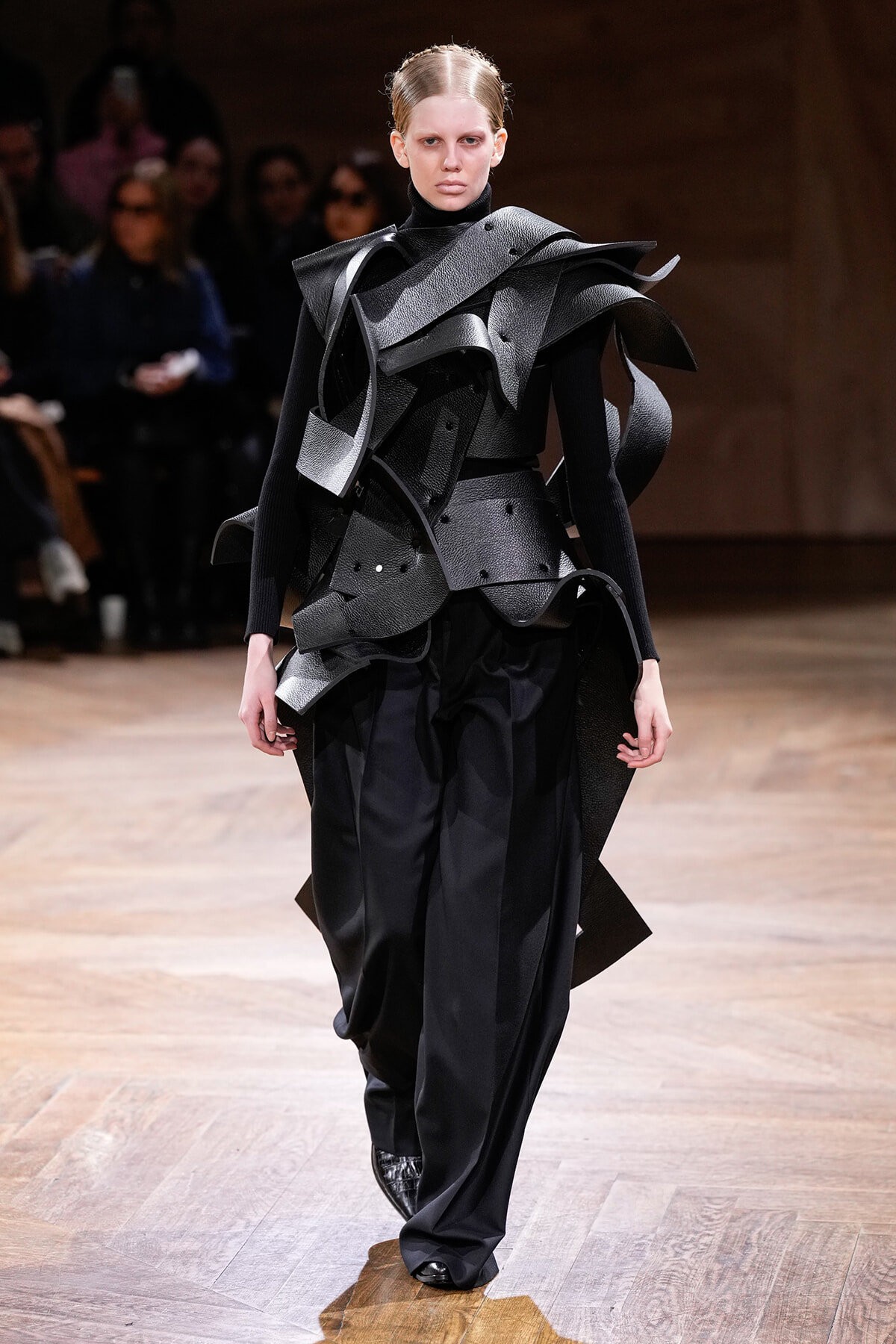 Model walking the runway in a futuristic black armor-inspired outfit with sculptural layered panels and oversized shoulders.