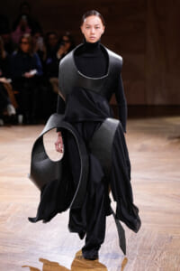 Model walks a fashion runway wearing an avant-garde black ensemble with large curved leather panels and flowing fabric.