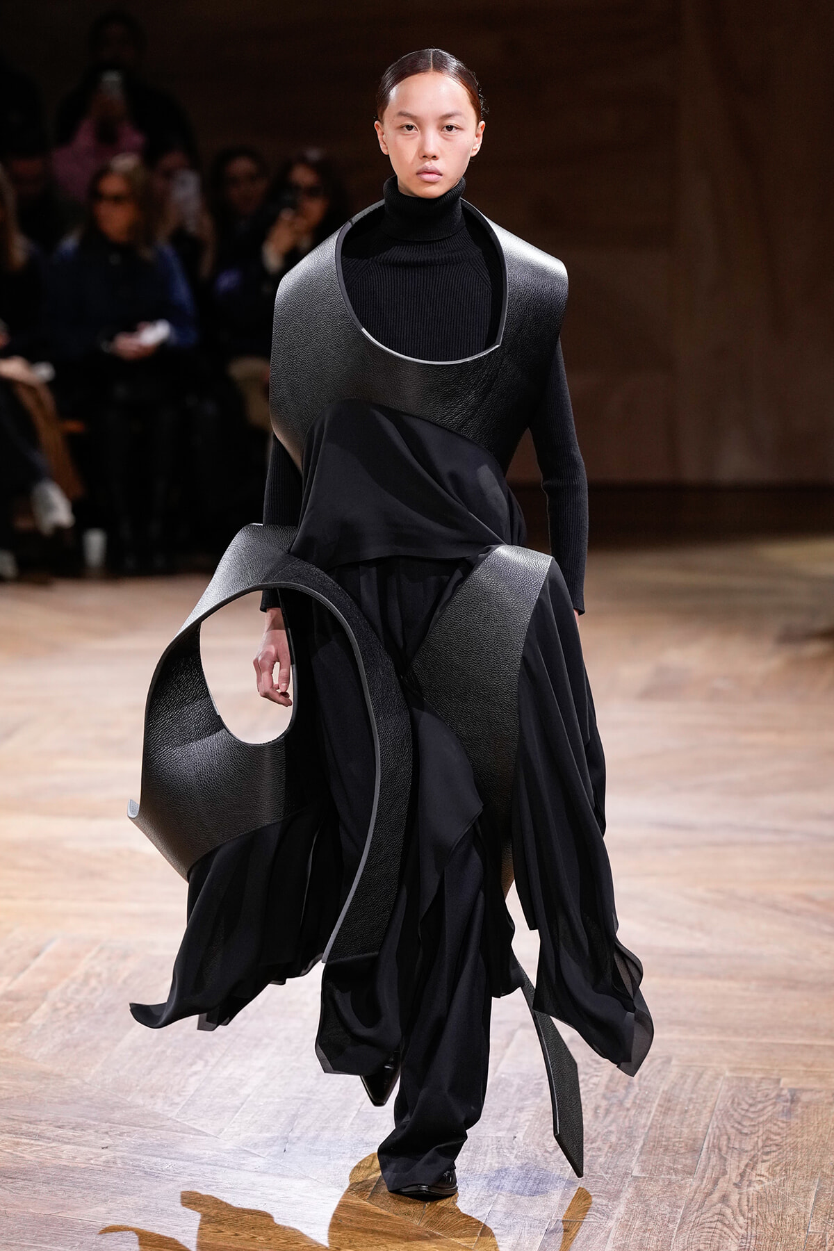 Model walks a fashion runway wearing an avant-garde black ensemble with large curved leather panels and flowing fabric.