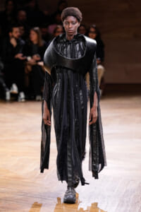 Model on a runway wearing a dramatic black sculptural top with curved shoulder armor and a flowing sheer skirted dress.