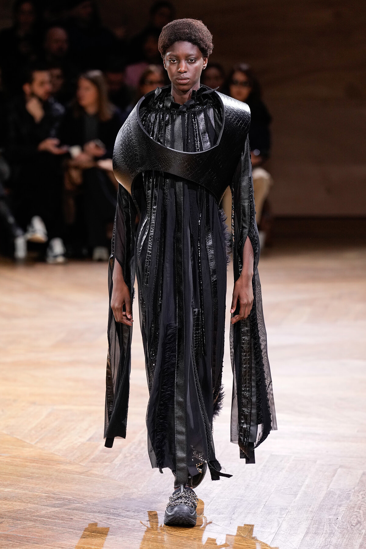 Model on a runway wearing a dramatic black sculptural top with curved shoulder armor and a flowing sheer skirted dress.