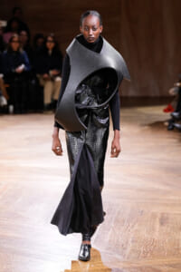 Runway model wearing a large curved gray sculptural neckpiece over a black outfit with a metallic-textured skirt.