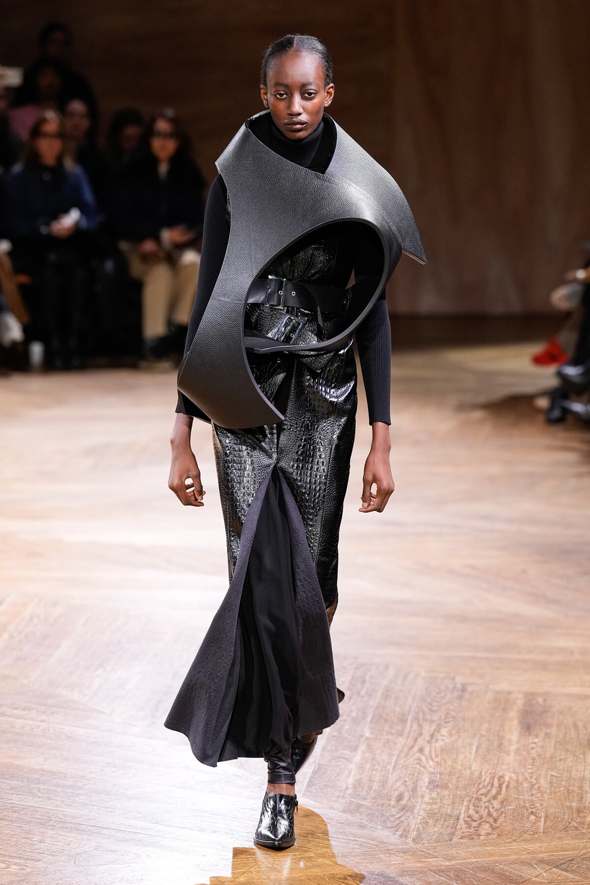 Runway model wearing a large curved gray sculptural neckpiece over a black outfit with a metallic-textured skirt.