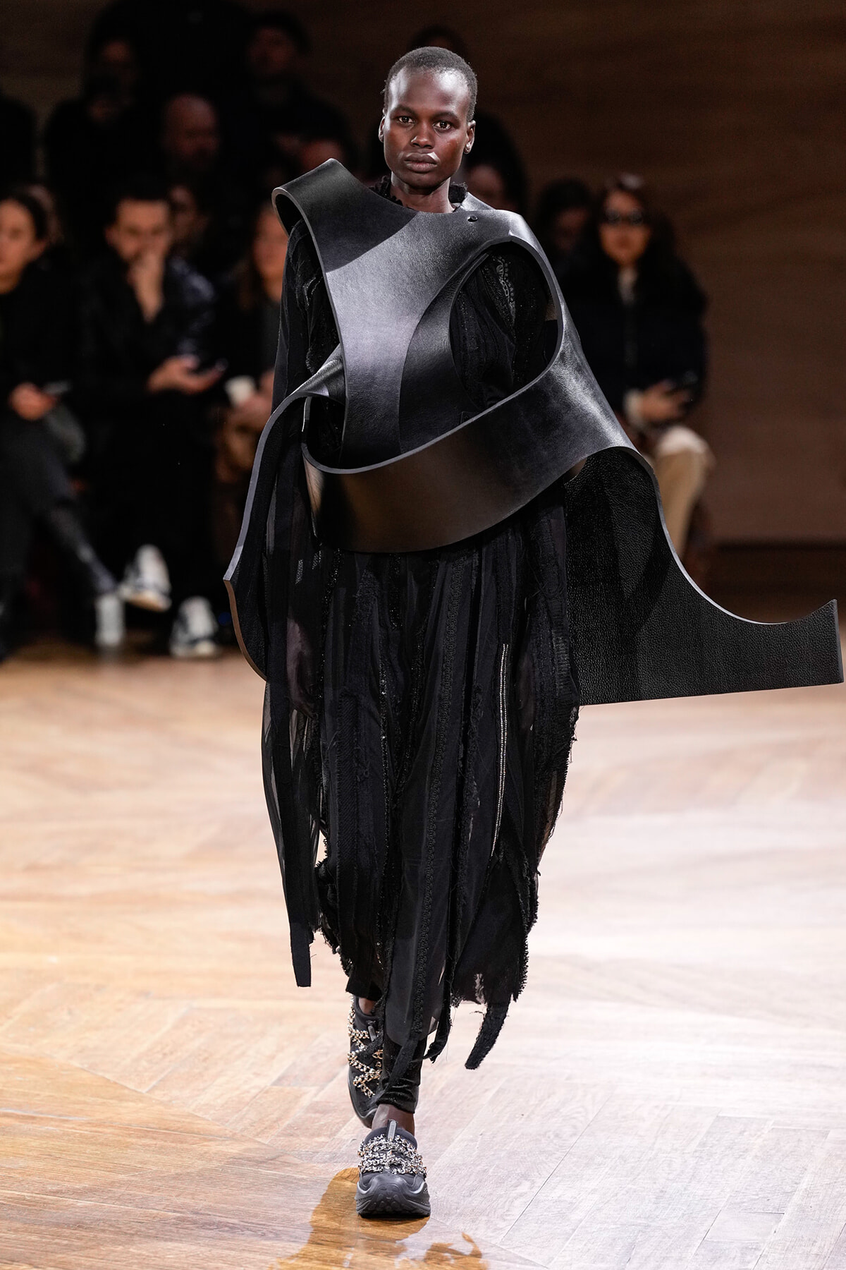 Model on a runway wearing an oversized black sculptural outfit with curved panels; audience blurred in background.