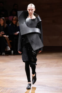 Model on a runway wearing an avant-garde black metallic outfit with oversized curved panels wrapping the torso and shoulder area.