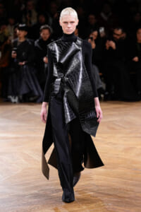 Model on a runway wearing a black turtleneck and an asymmetric, crocodile-embossed leather overlay with layered panels