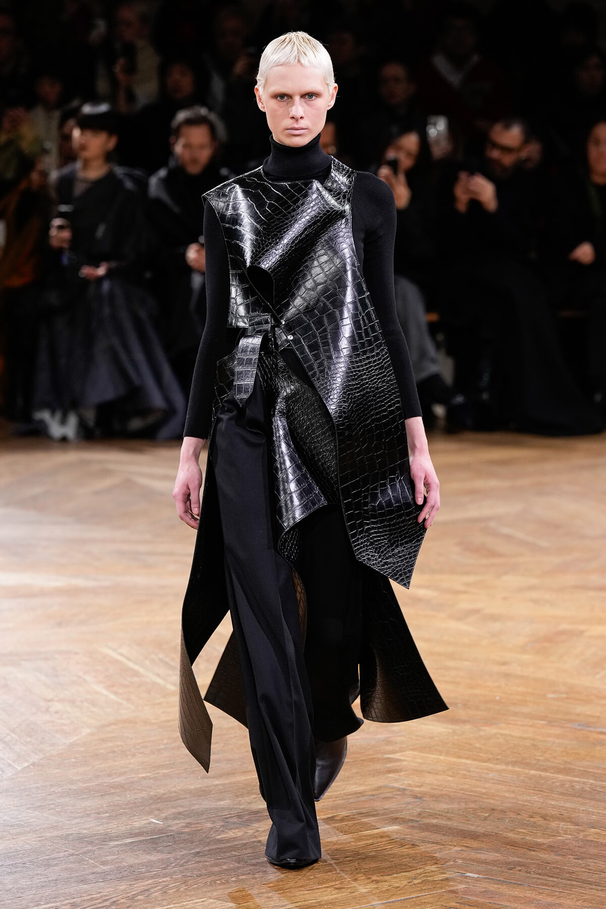 Model on a runway wearing a black turtleneck and an asymmetric, crocodile-embossed leather overlay with layered panels