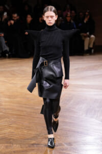 Model walks a runway in a black turtleneck with sculptural, winged shoulders and a layered black leather wrap skirt with a belt.