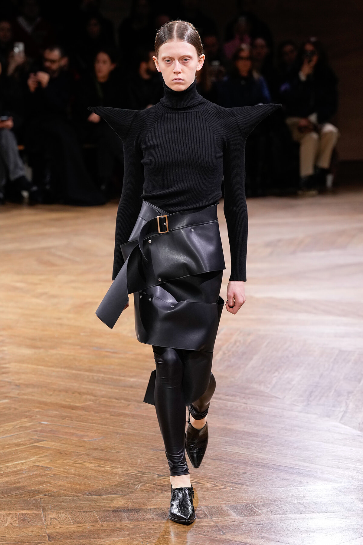 Model walks a runway in a black turtleneck with sculptural, winged shoulders and a layered black leather wrap skirt with a belt.