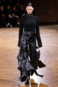 Female model walking a fashion runway in a black ribbed turtleneck and sculptural black leather strap skirt with multiple belts and metal accents.