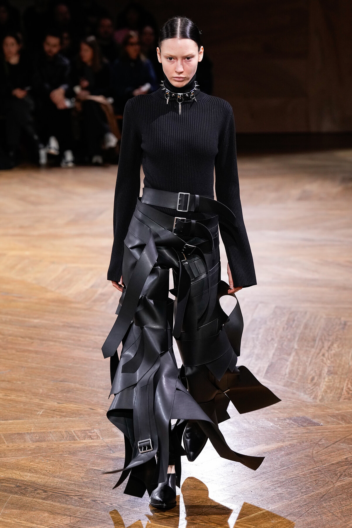 Female model walking a fashion runway in a black ribbed turtleneck and sculptural black leather strap skirt with multiple belts and metal accents.