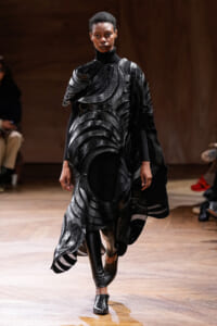 Model walks the runway in a black sculptural avant-garde outfit with layered curved panels, leather-like texture, and boots, during a fashion show.