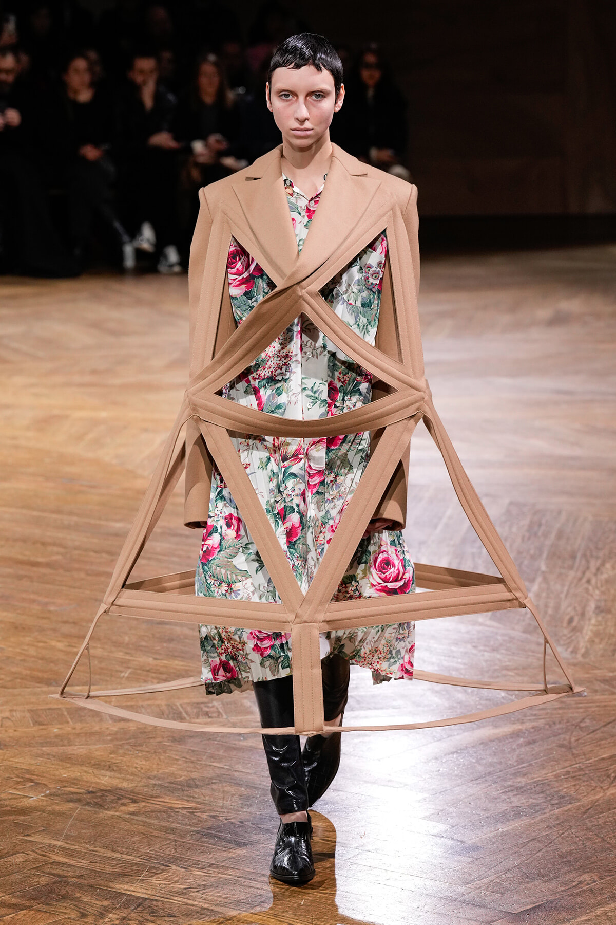 Model on a runway wearing a beige geometric harness over a floral dress; audience sits in the background.