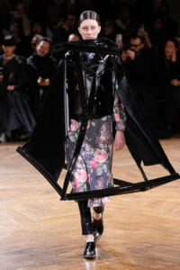 Model walking the runway in a floral dress with an oversized black plastic geometric frame around the shoulders, creating a sculptural silhouette.