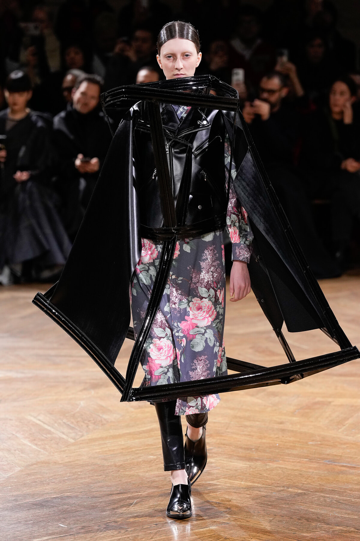 Model walking the runway in a floral dress with an oversized black plastic geometric frame around the shoulders, creating a sculptural silhouette.