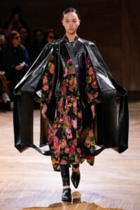 Model walks the runway in a dramatic black leather cape-style coat over a colorful floral dress, with pointed boots on a wooden catwalk.