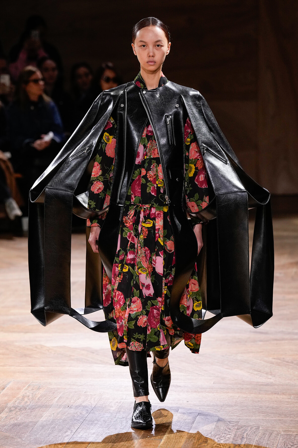 Model walks the runway in a dramatic black leather cape-style coat over a colorful floral dress, with pointed boots on a wooden catwalk.