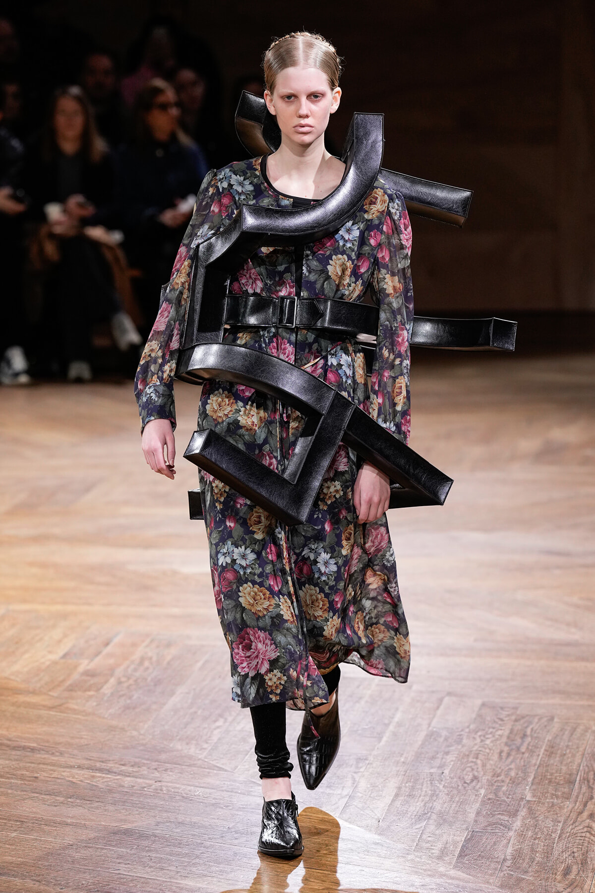 Model on a runway wearing a floral print dress with oversized black leather geometric wraps around the torso and shoulders.