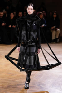 Model walking a runway in an avant-garde black outfit with large plastic frame structures around the torso and shoulders.