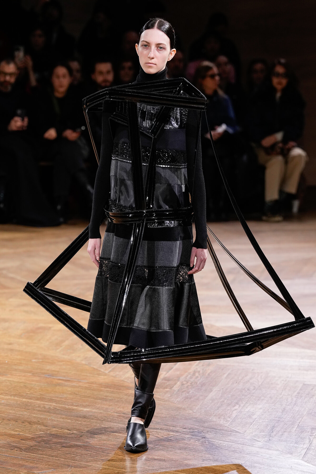 Model walking a runway in an avant-garde black outfit with large plastic frame structures around the torso and shoulders.