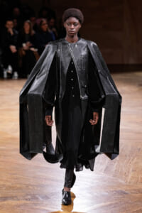 Model walking the runway in a metallic-black oversized cape with long draped panels and textured surface.