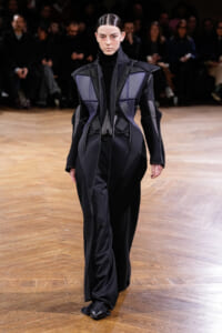 Model walking a fashion runway in a black and purple geometric, sculptural coat with structured panels.