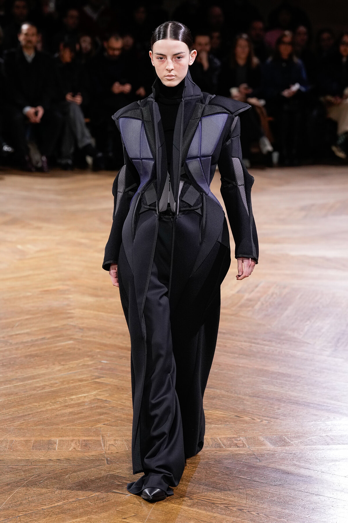 Model walking a fashion runway in a black and purple geometric, sculptural coat with structured panels.