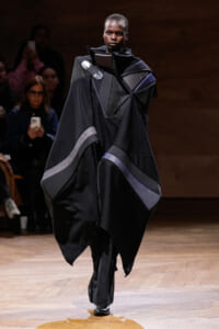 Model on a runway wearing a voluminous black garment with geometric panels and subtle purple accents.