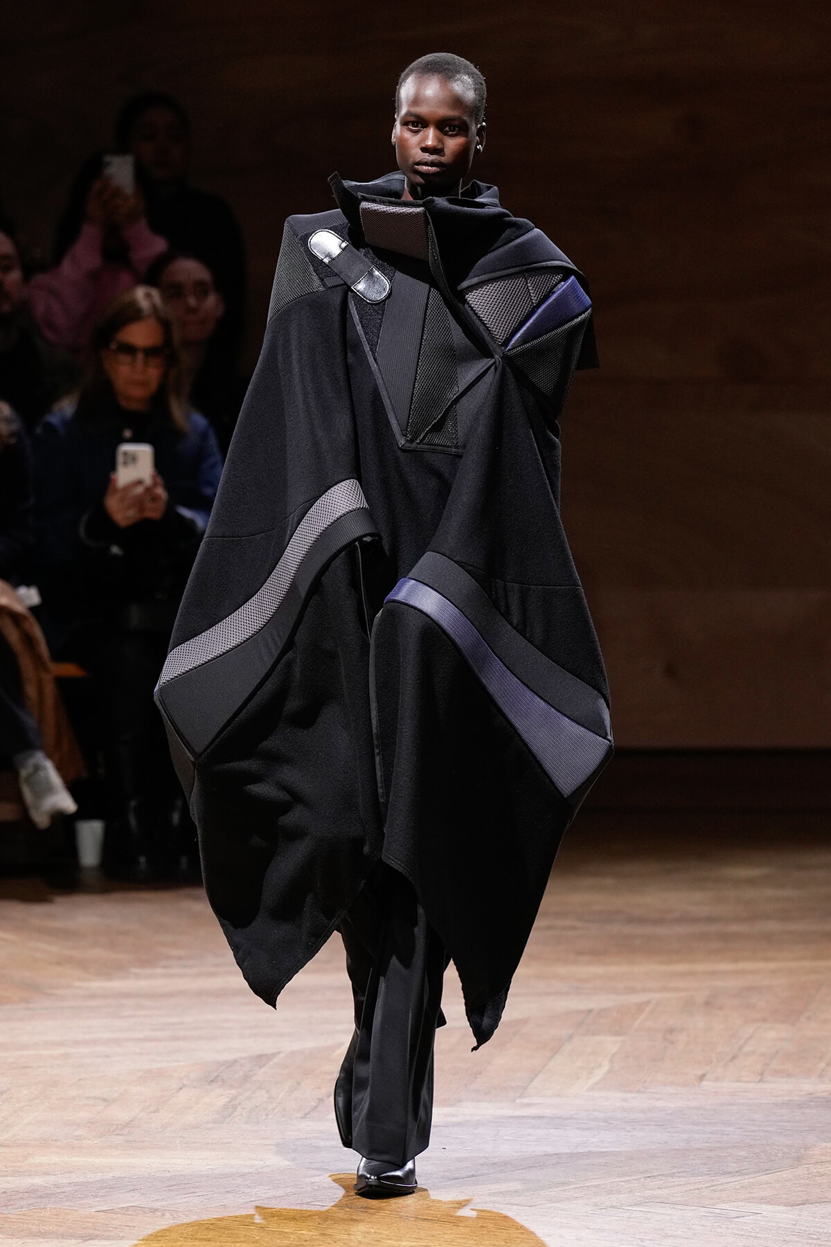 Model on a runway wearing a voluminous black garment with geometric panels and subtle purple accents.