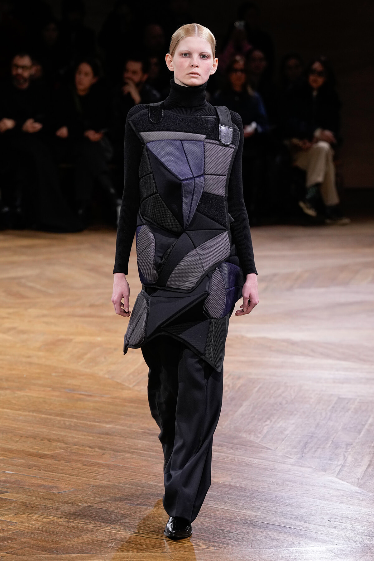 Model walking a runway in a black turtleneck with a sculptural, geometric patchwork dress in dark grays and purples.