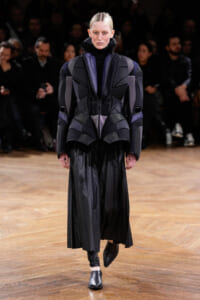 Model walking a runway in a dark, geometric padded coat with purple accents