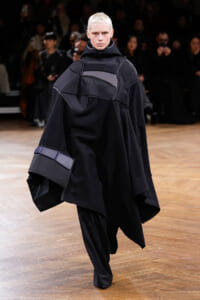 Model walking a runway in an oversized black avant-garde outfit with layered geometric panels, high collar hood, and wide sleeves; dark, minimal styling with a light background audience.